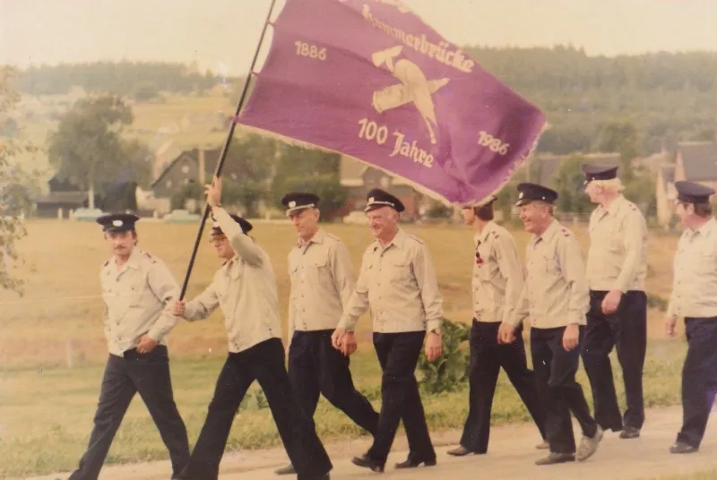 Farbfoto: Feuerwehrmänner marschieren im Festumzug, vorneweg die violette Fahne mit der Aufschrift 100 Jahre 1886–1986