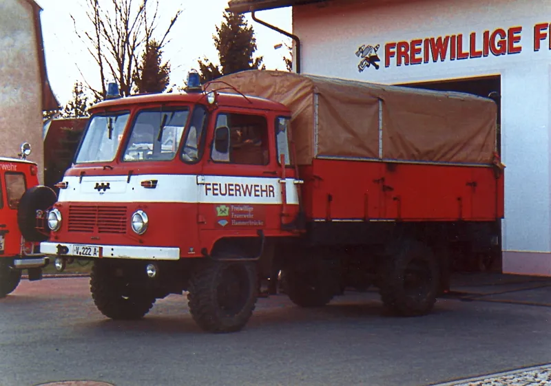 Farbfoto: der rot-weiße Robur LO mit Plane vor dem Gerätehaus mit Schriftzug Freiwillige Feuerwehr